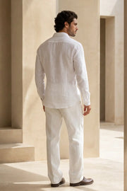 A man wearing a white long-sleeve linen shirt and matching white trousers, shown from the back in a sunlit architectural setting with beige columns.