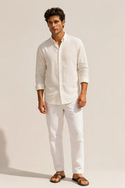 A man modeling a white long-sleeve linen button-down shirt and matching white trousers with brown leather sandals, standing against a warm beige studio background.