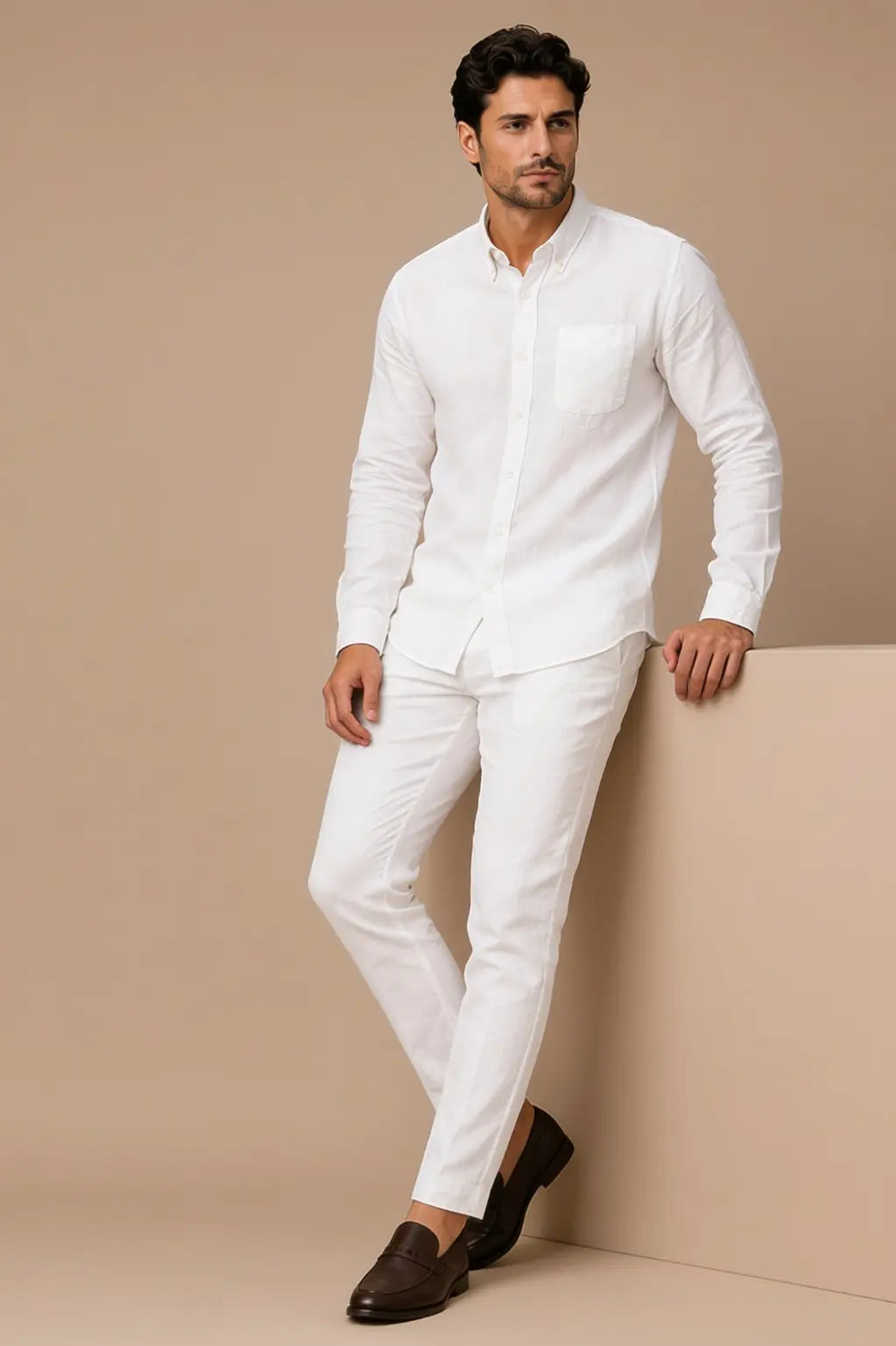 A man modeling a white long-sleeve linen shirt and matching white trousers, paired with dark brown loafers, standing against a beige studio backdrop.