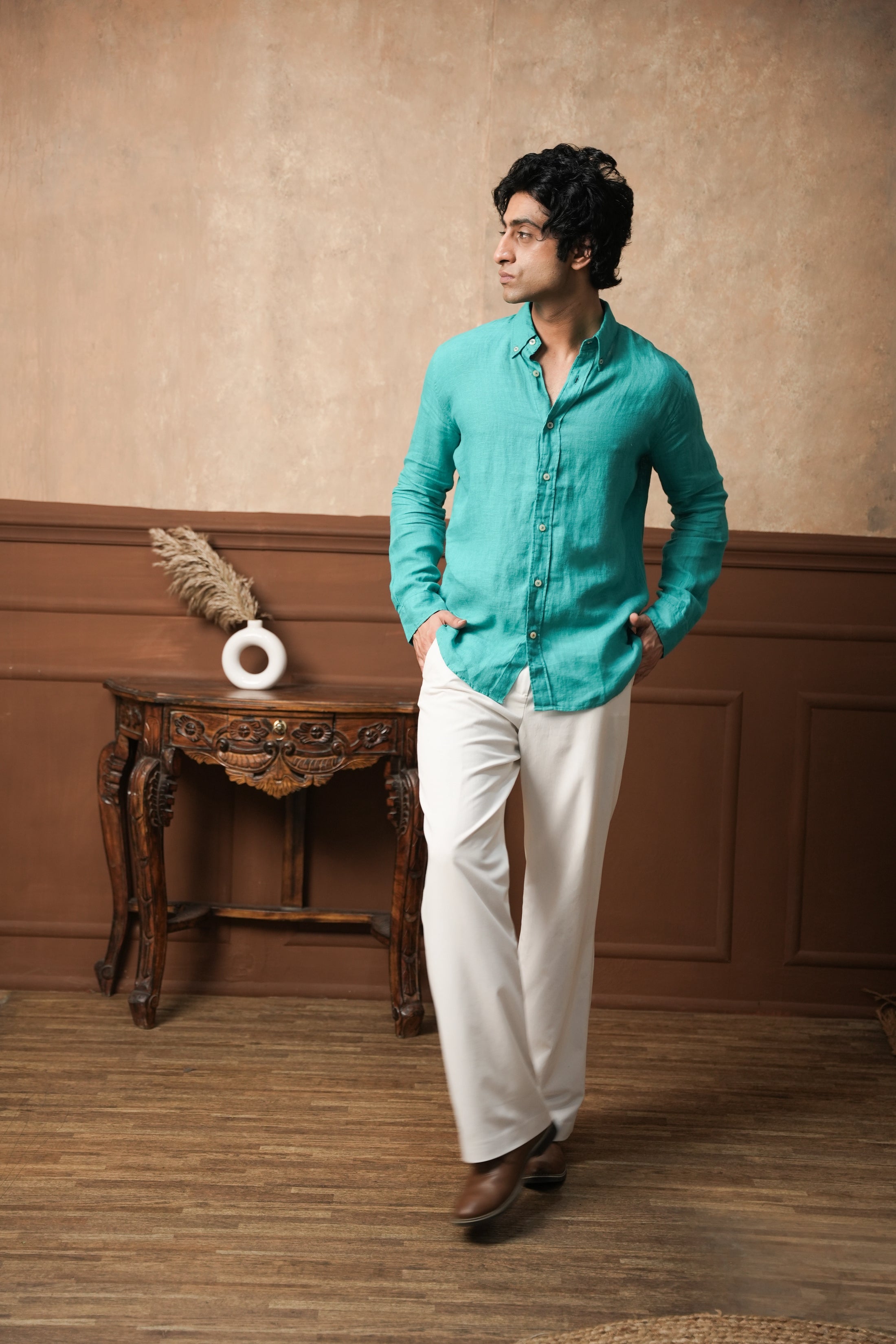 Man in a teal shirt and white pants standing in a room with wooden flooring and a decorative table.